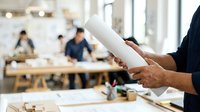 Mature male professional holding rolled architectural plans in modern office workspace with colleagues collaborating in background, suitable for business, construction, and corporate projects.