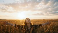 Young woman with arms outstretched celebrating freedom in golden wheat field at sunset, expressing joy and gratitude in warm sunlight.