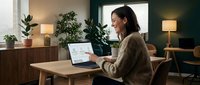 Young Asian woman working on laptop in modern cozy home office with plants and warm lighting, remote work lifestyle and productivity concept.