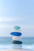Balanced stack of smooth sea glass stones in blue, white and green colors on sandy beach with ocean backdrop for wellness and meditation concepts.