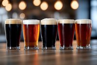 Variety of craft beer glasses lined up on wooden bar counter showing different styles from dark stout to amber ale with blurred pub lights background.