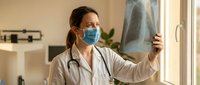 Female doctor in white coat and medical mask examining chest x-ray film by window in modern clinic office for patient diagnosis and healthcare consultation.