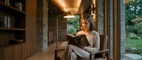 Young blonde woman reading book in modern luxury home library with warm lighting and garden view creating peaceful atmosphere for relaxation and learning.