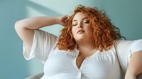 Young curly-haired woman with red hair relaxing with eyes closed in white top against blue background. Portrait of confident plus-size female model posing.