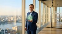Professional middle-aged Caucasian businessman in navy suit holding glowing green recycling symbol in modern office with city skyline view. Sustainability concept.
