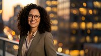 Professional woman with curly dark hair and glasses wearing business blazer smiling at camera with blurred city skyline and golden lights in background at dusk.