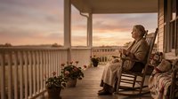Elderly woman relaxing in rocking chair on wooden porch during golden hour sunset, peaceful retirement lifestyle and home comfort concept.