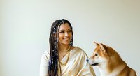 Young woman with braided hair and warm brown skin tone wearing a gold satin shirt, smiling while posing with a Shiba Inu dog against neutral background for portrait use.