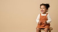 Happy toddler girl with dark curly hair, mixed ethnicity, wearing orange overalls and white long-sleeve shirt, smiling and laughing against neutral beige background for childcare, parenting, and family content.