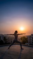Woman practicing yoga warrior pose on rooftop at sunrise with urban cityscape background. Fitness wellness meditation concept for healthy lifestyle.