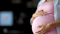 Pregnant woman in pink dress tenderly holding her belly against dark background, showing maternal care and connection with unborn baby during pregnancy.