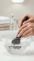 Woman hands cleaning makeup brush in glass bowl with soapy water in bright bathroom setting for beauty routine and cosmetic hygiene maintenance.