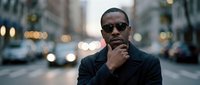 Thoughtful African American male professional wearing sunglasses and dark blazer on urban city street with blurred traffic and buildings in background.