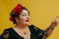 Hispanic plus size woman with red roses in hair, red lipstick, face gems, and tattoos smiling and pointing against yellow background. Modern body positive portrait.