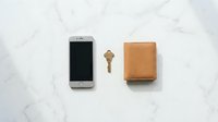 Smartphone with brown leather wallet and brass key arranged on white marble surface for minimalist lifestyle and everyday carry concepts.
