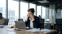 Stressed businesswoman holding head in hands at modern office desk with laptop, experiencing work pressure and mental fatigue in corporate environment.
