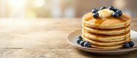 Stack of golden pancakes topped with fresh blueberries and honey drizzle on white plate, wooden table background with warm sunlight, breakfast and brunch food photography.