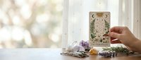 Hand holding ornate tarot card with mystical symbols near window with crystals and spiritual items for divination reading session.