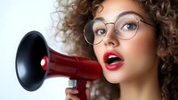 Young woman with curly brown hair and fair complexion wearing round glasses holds red megaphone to mouth against white background. Suitable for announcements, marketing campaigns, communication concepts.