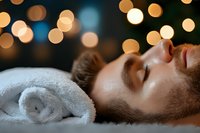 Young Caucasian man with beard relaxing during spa treatment with soft bokeh lights in background, lying on massage table with white towel.