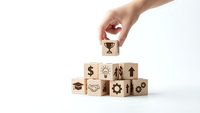 Hand placing trophy cube on wooden blocks with business icons representing success achievement and goal completion on white background.
