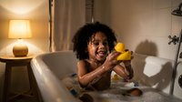 Happy African American toddler girl playing with yellow rubber duck in bathtub during evening bath time routine at home.