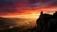 Silhouette of hiker standing on rocky cliff edge at dramatic sunset with golden and orange sky over mountain valley landscape below.