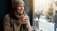 Young woman in cozy knit sweater and beanie enjoying hot drink by window during winter holiday season with festive street decorations in background.