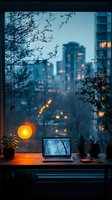 Cozy workspace with laptop on desk by window overlooking city lights at dusk, warm lamp glow contrasts with blue twilight cityscape, houseplants frame peaceful scene.