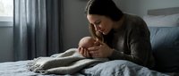 Young mother tenderly holding and kissing newborn baby while sitting on bed in cozy bedroom. Intimate bonding moment between parent and infant in soft natural light.