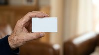 Close-up of mature male hand holding blank white business card with blurred interior background, suitable for corporate branding and professional networking presentations.