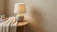 Wooden table lamp with linen shade next to folded cream textile on round wooden side table in warm minimalist interior setting.