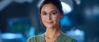 Young Asian female medical professional wearing glasses and green scrubs smiling warmly at camera against blurred hospital background.