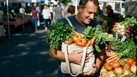 Middle-aged Caucasian man holding canvas bag filled with fresh organic vegetables at bustling farmers market with shoppers browsing produce stands.