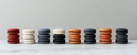 Colorful macarons arranged in neat stacks on white marble surface, showing variety of flavors in red, white, gray and orange tones against neutral background.