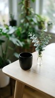 Minimalist wooden table with black ceramic mug and eucalyptus branch in glass vase, cozy home interior with natural light and green plants in background.