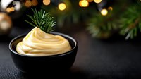Creamy yellow sauce swirled in black bowl garnished with fresh rosemary sprig against dark background with golden bokeh lights and pine branches.
