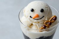 Creamy vanilla ice cream dessert shaped like a snowman with chocolate eyes, carrot nose, and pretzel arms in a glass cup on light background.