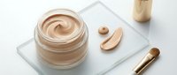Luxury beige foundation cream in glass jar with makeup brush and cosmetic applicator on white marble surface for skincare and beauty routine.
