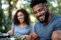 Happy African American man laughing while cooking outdoors with woman in background during summer barbecue gathering in natural park setting.