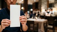 Asian male professional in dark business attire holding blank white paper card in corporate event venue with blurred colleagues and elegant interior background for mockup and branding purposes.