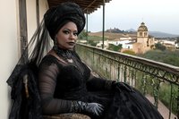 African woman in elegant Victorian black dress with ornate headwrap sitting on historic balcony overlooking Mediterranean village architecture.