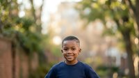 Young African American boy, approximately 6-8 years old, wearing a dark blue t-shirt, smiling at camera in tree-lined residential street setting with soft natural lighting.