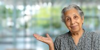 Senior woman with gray hair gesturing with open palm against blurred bright background, representing wisdom, hospitality, or presentation concept.