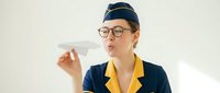 Young woman, Caucasian ethnicity, wearing airline stewardess uniform with navy blue jacket and yellow collar details, glasses and captain's hat, holding paper airplane. Flight attendant concept.