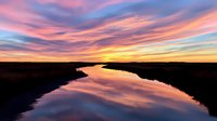 Dramatic sunset sky with vibrant pink and orange clouds reflected in calm river water creating mirror effect across peaceful landscape horizon.