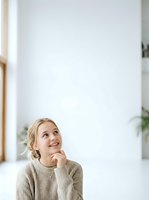 Young woman with blonde hair in casual beige sweater, hand to chin in thoughtful pose, gazing upward in bright minimalist interior space for contemplation and decision-making concepts.