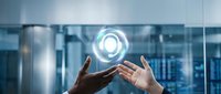 Two hands holding glowing circular technology icon above modern office building interior with glass windows and cityscape background for business innovation concepts.