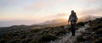 Hiker with backpack walking on mountain trail at sunrise with misty peaks in background, adventure and outdoor exploration.