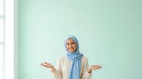 Woman wearing light blue hijab and beige cardigan gestures with open palms against mint green wall, expressing uncertainty or welcome gesture for lifestyle and diversity content.
